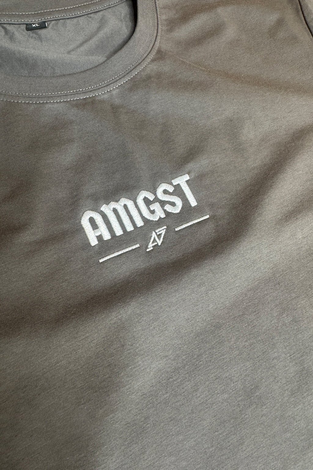 A close-up of a heather gray t-shirt featuring the word 'AMGST' and a stylised 'AMGST' logo printed in white on the chest.